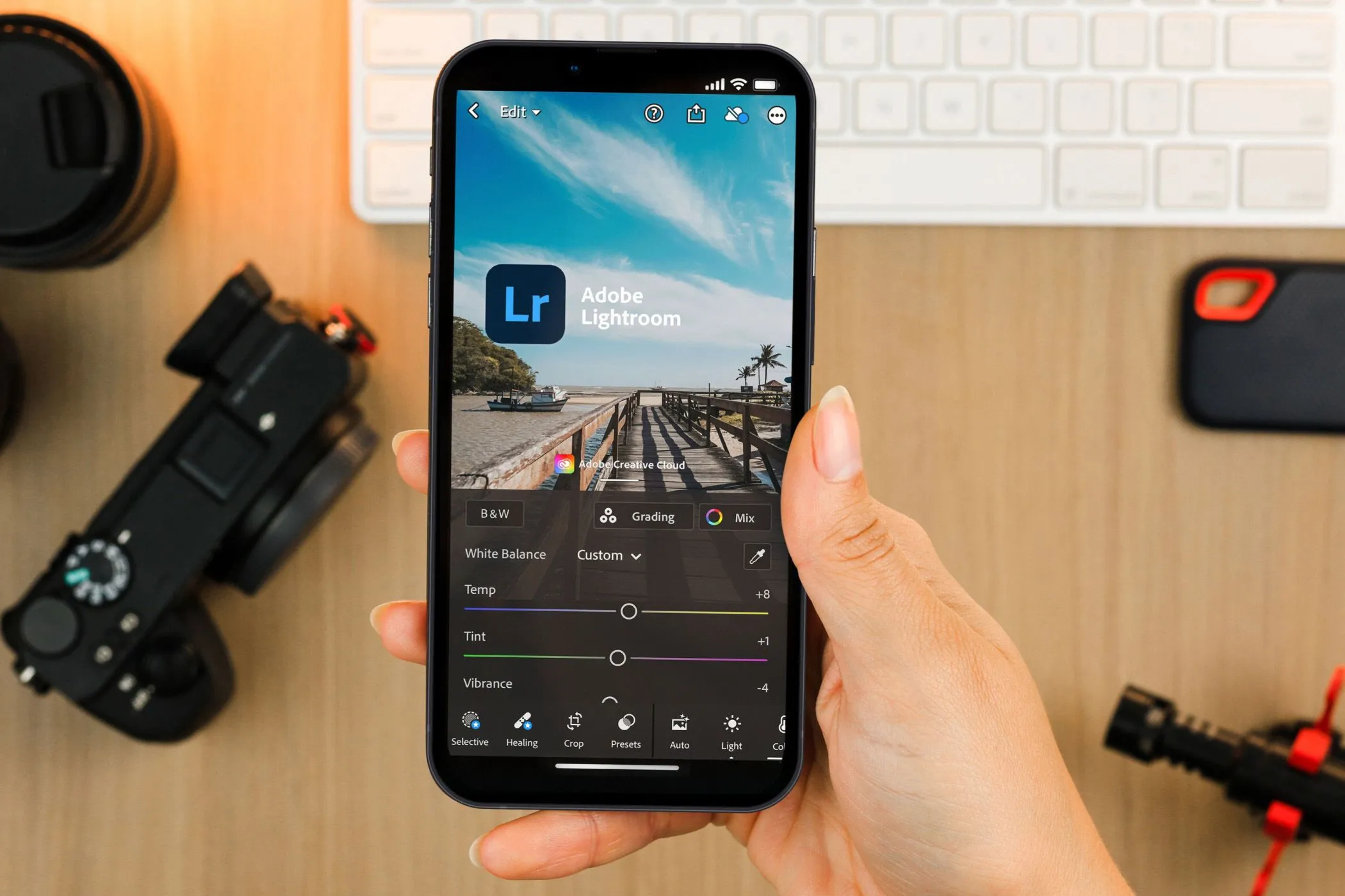 Bàn tay đang cầm điện thoại hiển thị ứng dụng Lightroom Mobile, với thiết bị máy ảnh chuyên nghiệp ở hậu cảnh, nhấn mạnh vai trò chỉnh sửa ảnh di động.