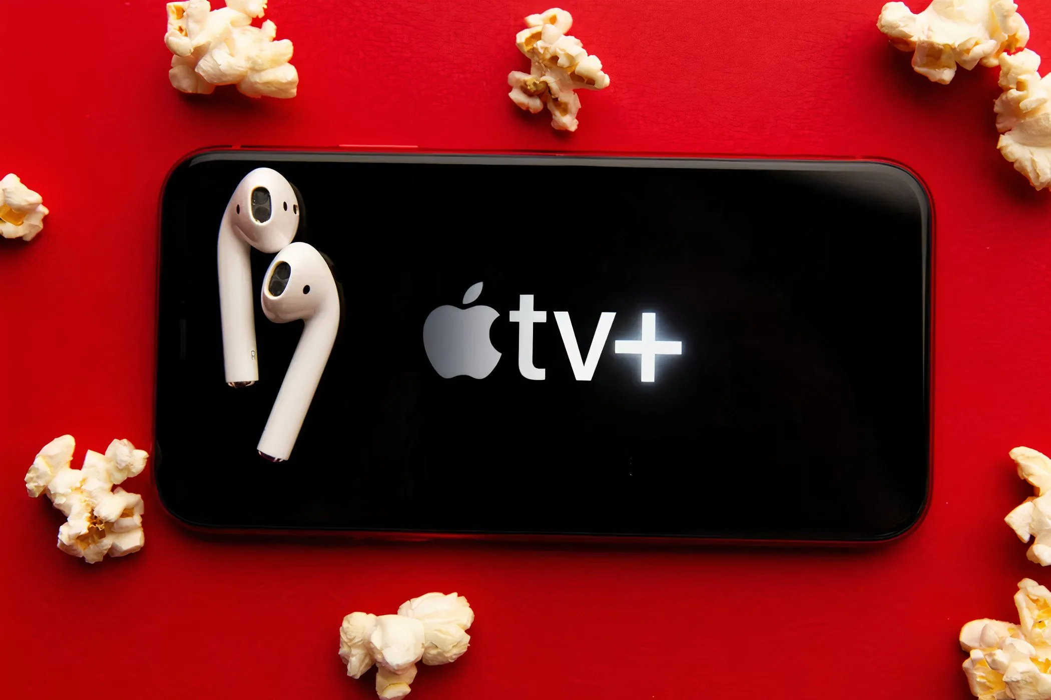 Hình ảnh logo Apple TV+ trên màn hình điện thoại thông minh, bên cạnh là bỏng ngô và tai nghe không dây, gợi ý trải nghiệm xem phim giải trí trên thiết bị di động