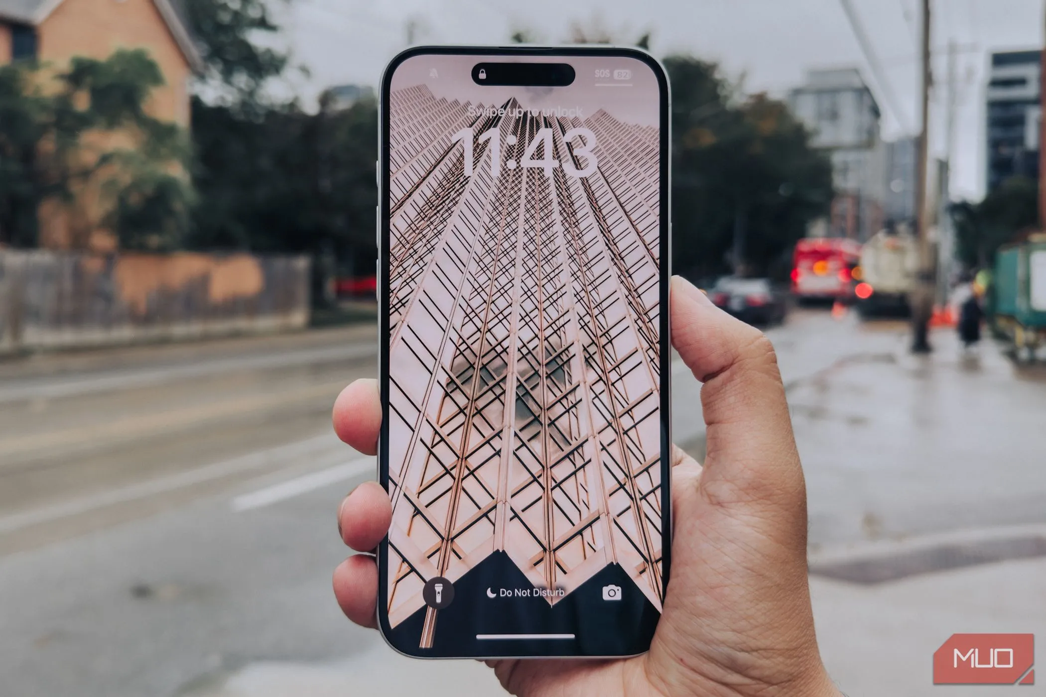 Màn hình iPhone 16 Plus với viền mỏng đang được cầm trên tay trước phông nền thành phố.