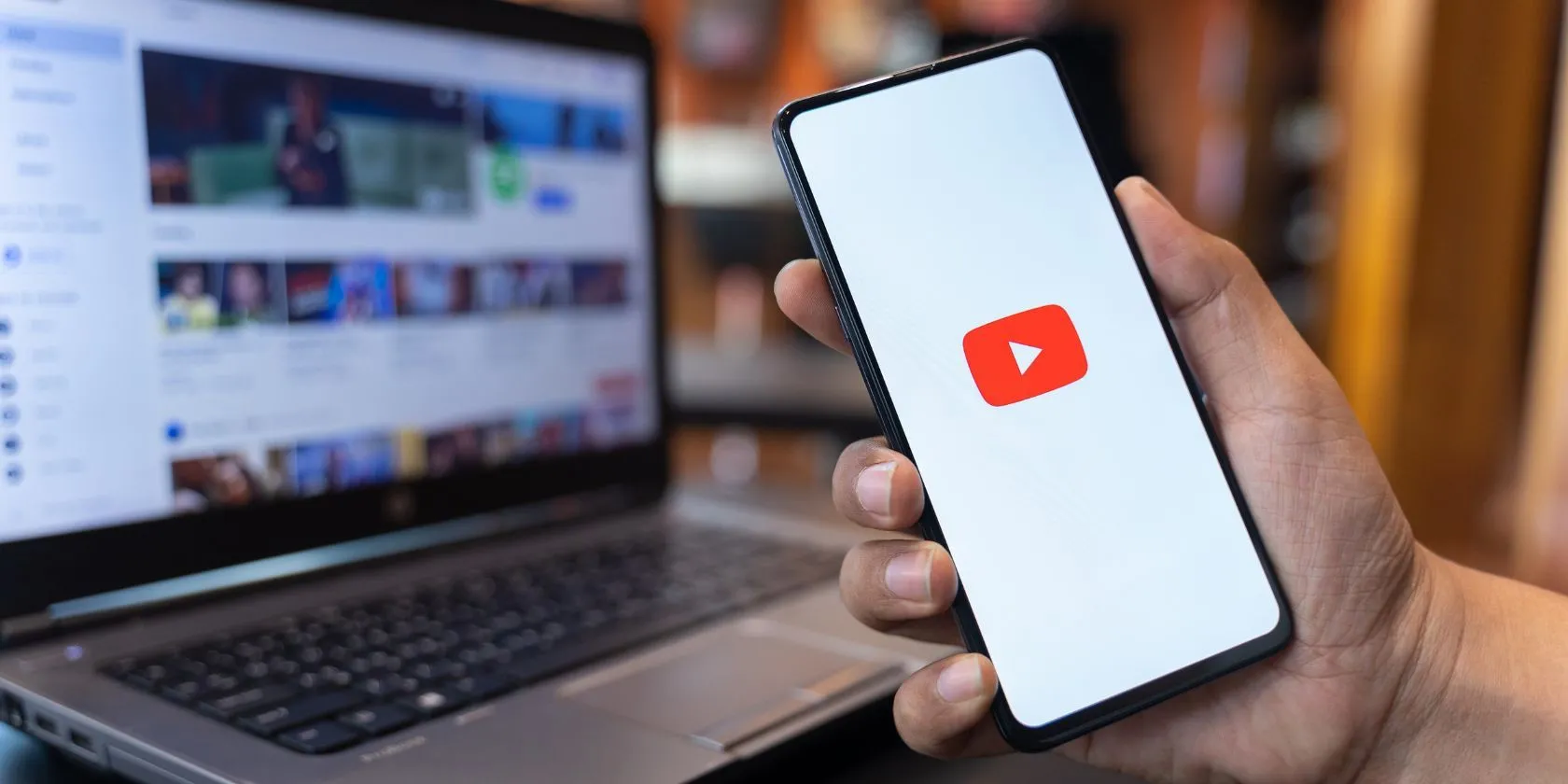 Một người đang cầm điện thoại di động mở ứng dụng YouTube, với trang web YouTube hiển thị trên màn hình máy tính phía sau, tượng trưng cho việc người dùng cần cảnh giác khi duyệt nội dung trên nền tảng này.