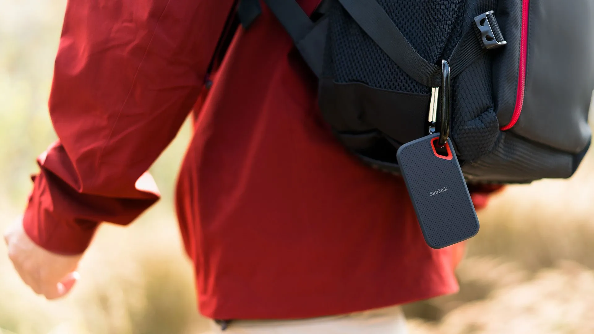 Ổ SSD di động SanDisk Extreme 8TB màu đen được móc vào balo của người đàn ông mặc áo khoác đỏ, minh họa khả năng di chuyển linh hoạt và dễ dàng mang theo.